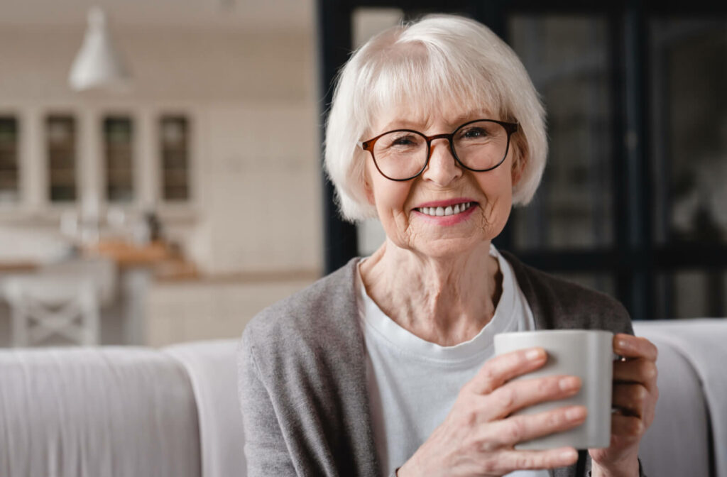 A senior woman with short, white hair and glasses sitting, smiling, and holding a cup of tea.