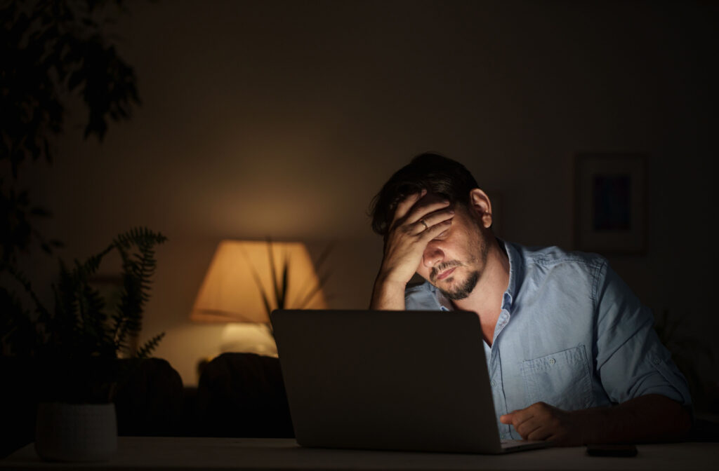 Man using a laptop in a dark room with his hand on his forehead, looking stressed