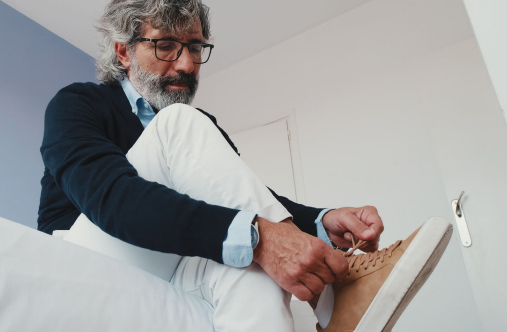 A styling senior man in a white room tying the lace of his right shoe.