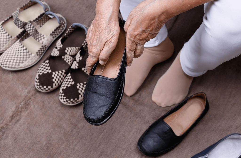 A senior woman with three pairs of shoes near her about to put on a laceless slip-on shoe on her right foot.