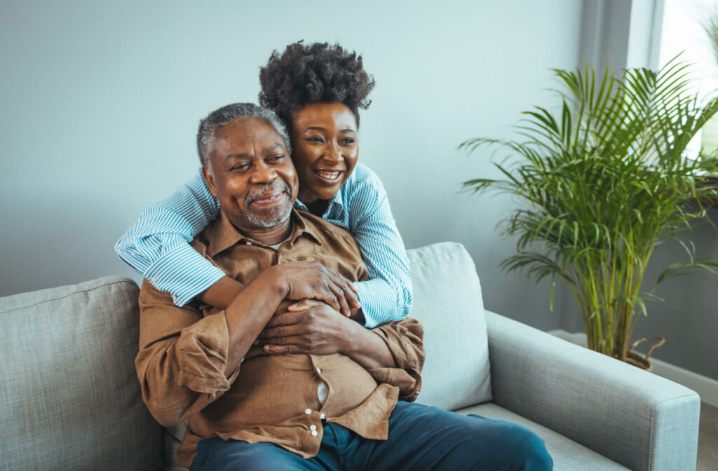 A woman hugging her senior father from behind while he is sitting on a couch.