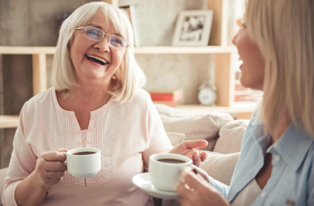 An older adult woman and her daughter sitting on a couch smiling and talking to each other while holding a cup of tea